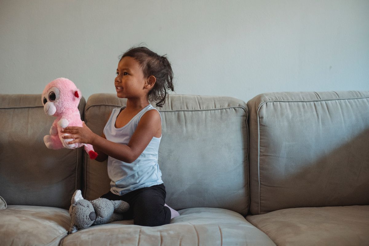 Adorable little Hispanic girl playing with soft toys on comfortable sofa at home