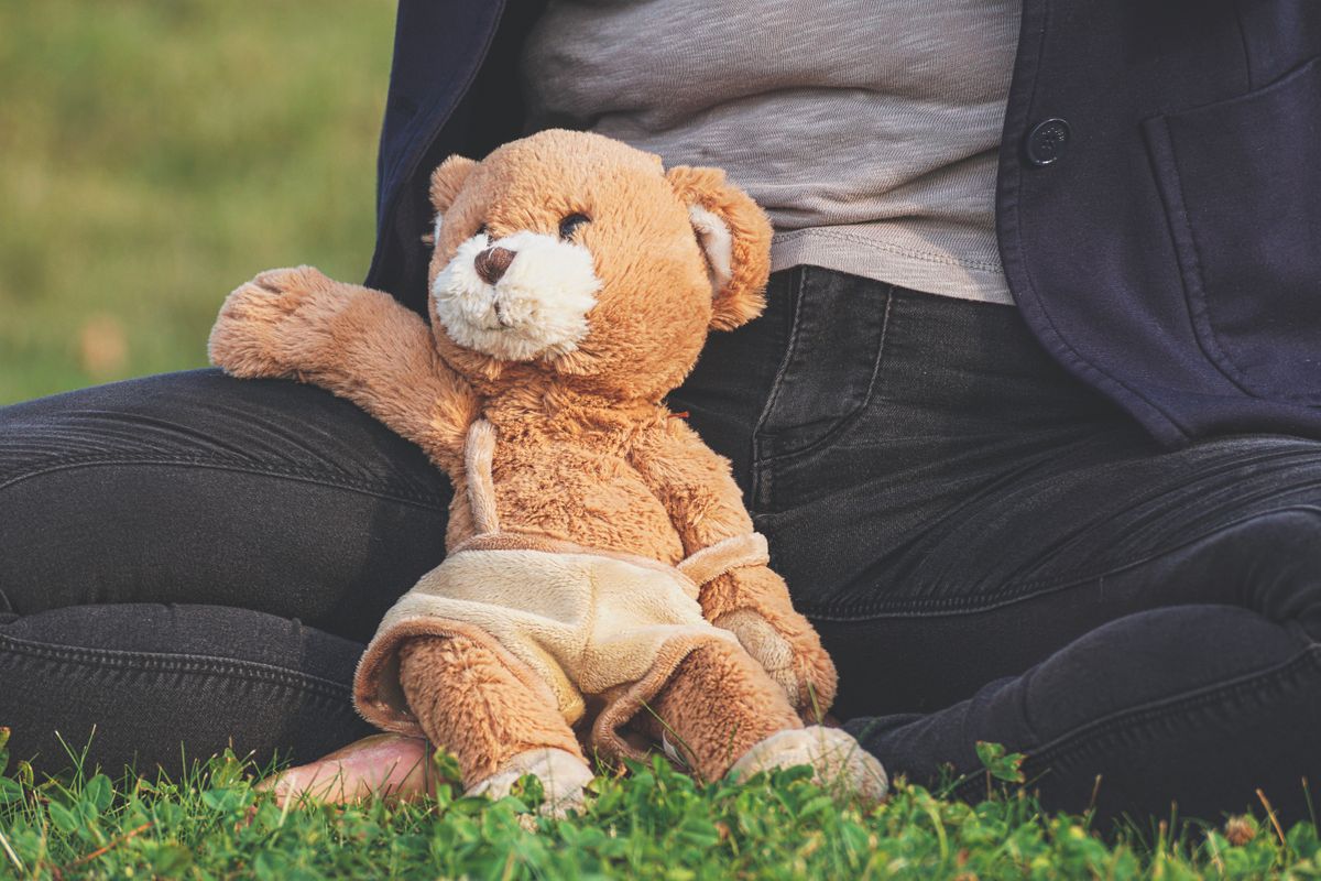 A plush teddy bear sits comfortably on a person's lap in an outdoor grassy environment, embodying warmth and comfort.