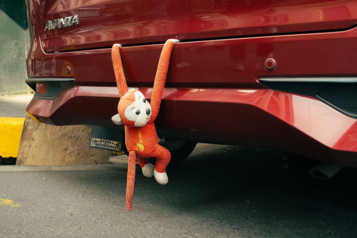 Playful stuffed monkey toy hanging from a red car bumper in an outdoor setting.
