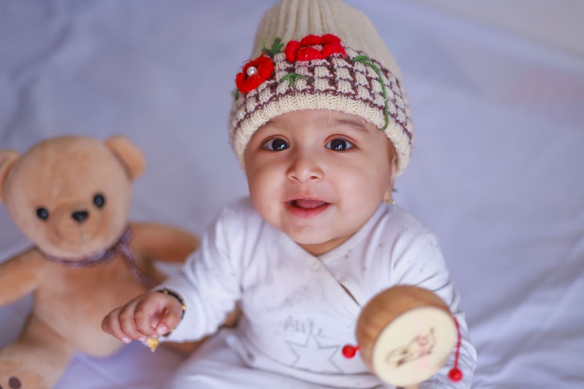 Cute baby smiling with a knitted cap, holding a wooden toy, and sitting beside a plush teddy bear.