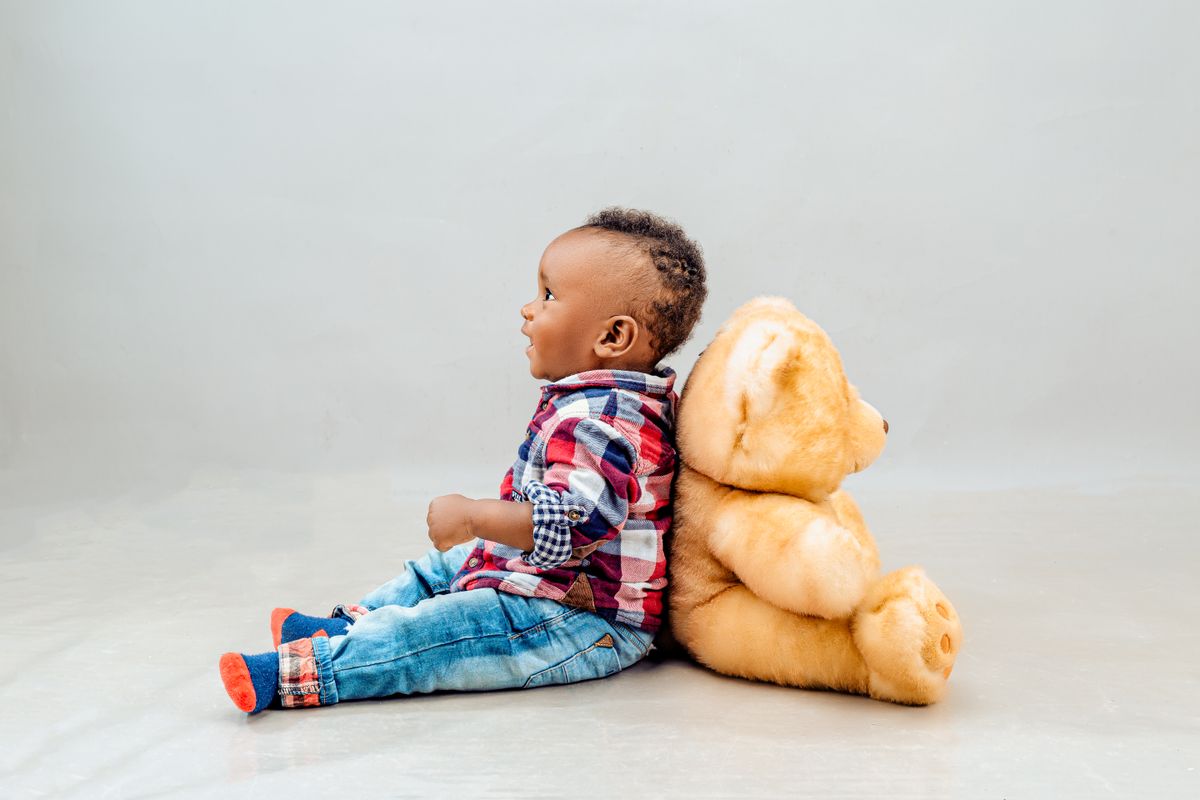 Adorable baby in plaid shirt sitting back-to-back with teddy bear, studio setting.