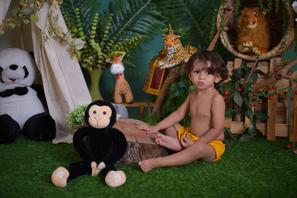 A young child in a jungle-themed room surrounded by stuffed animals and plants, sitting on fake grass.