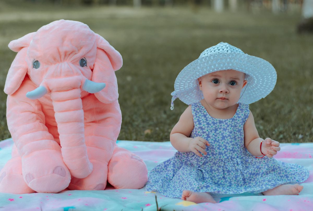 Cute baby girl in floral dress and hat sitting on a blanket with pink plush elephant outdoors.