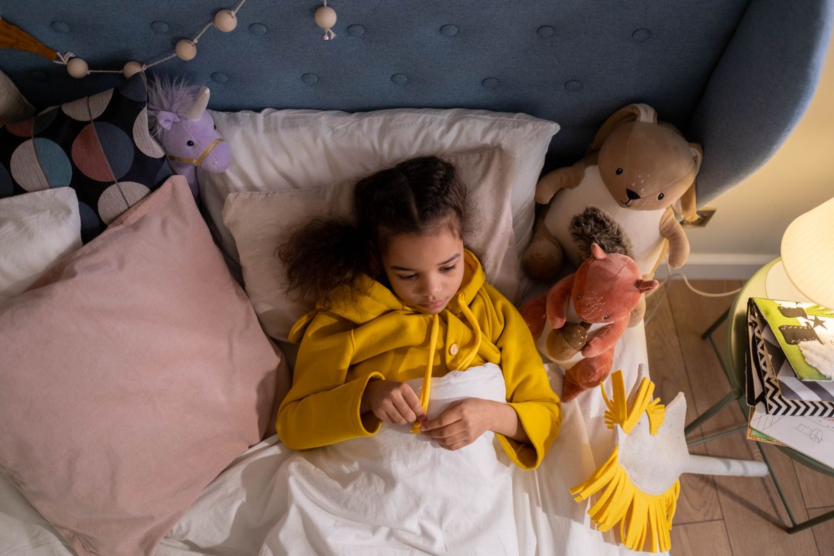 A young girl wearing a yellow hoodie lying in bed surrounded by plush toys, provides a cozy atmosphere.