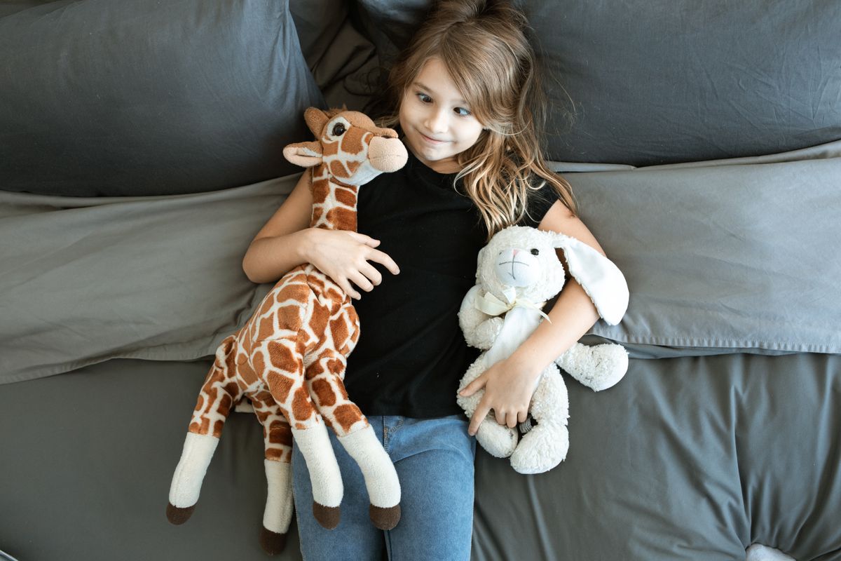 Young girl relaxing on bed, hugging giraffe and bunny plush toys, creating a cozy and playful atmosphere.