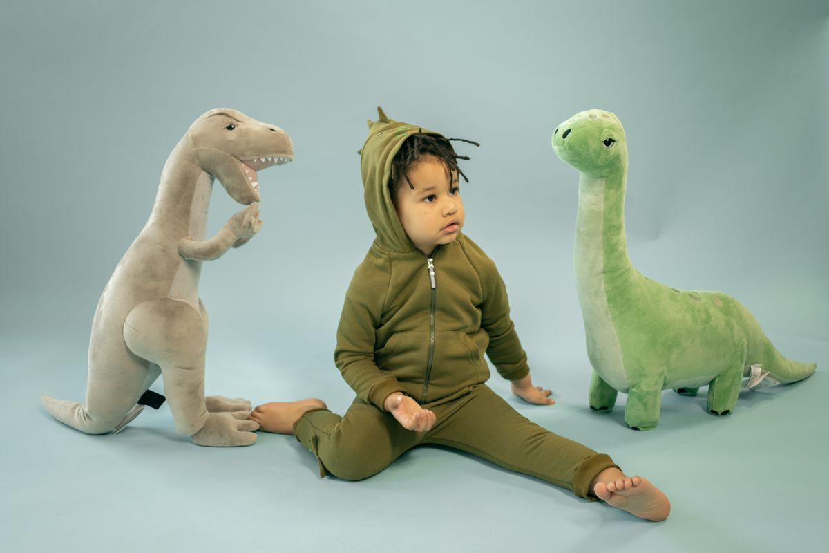 Full body of adorable barefoot African American boy in dinosaur wear sitting on light background near plush dinosaurs toys in studio
