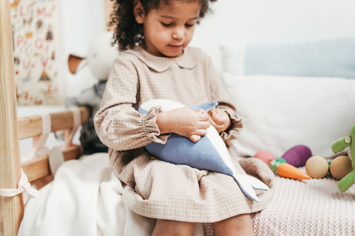 A young girl in a brown dress holds a whale plush toy, surrounded by colorful stuffed animals in a cozy indoor setting.