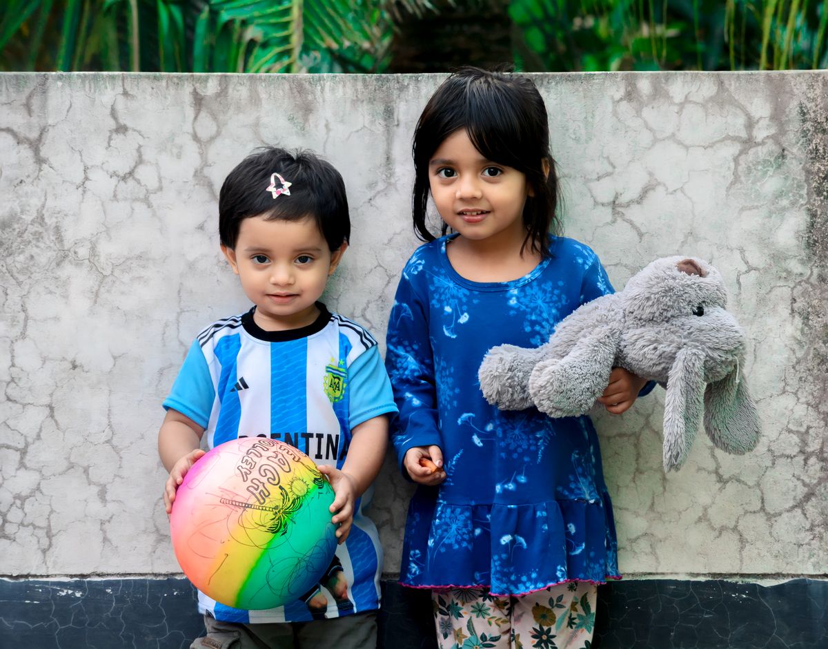 Two young children posing outdoors holding a colorful ball and plush toy, expressing innocence and joy.
