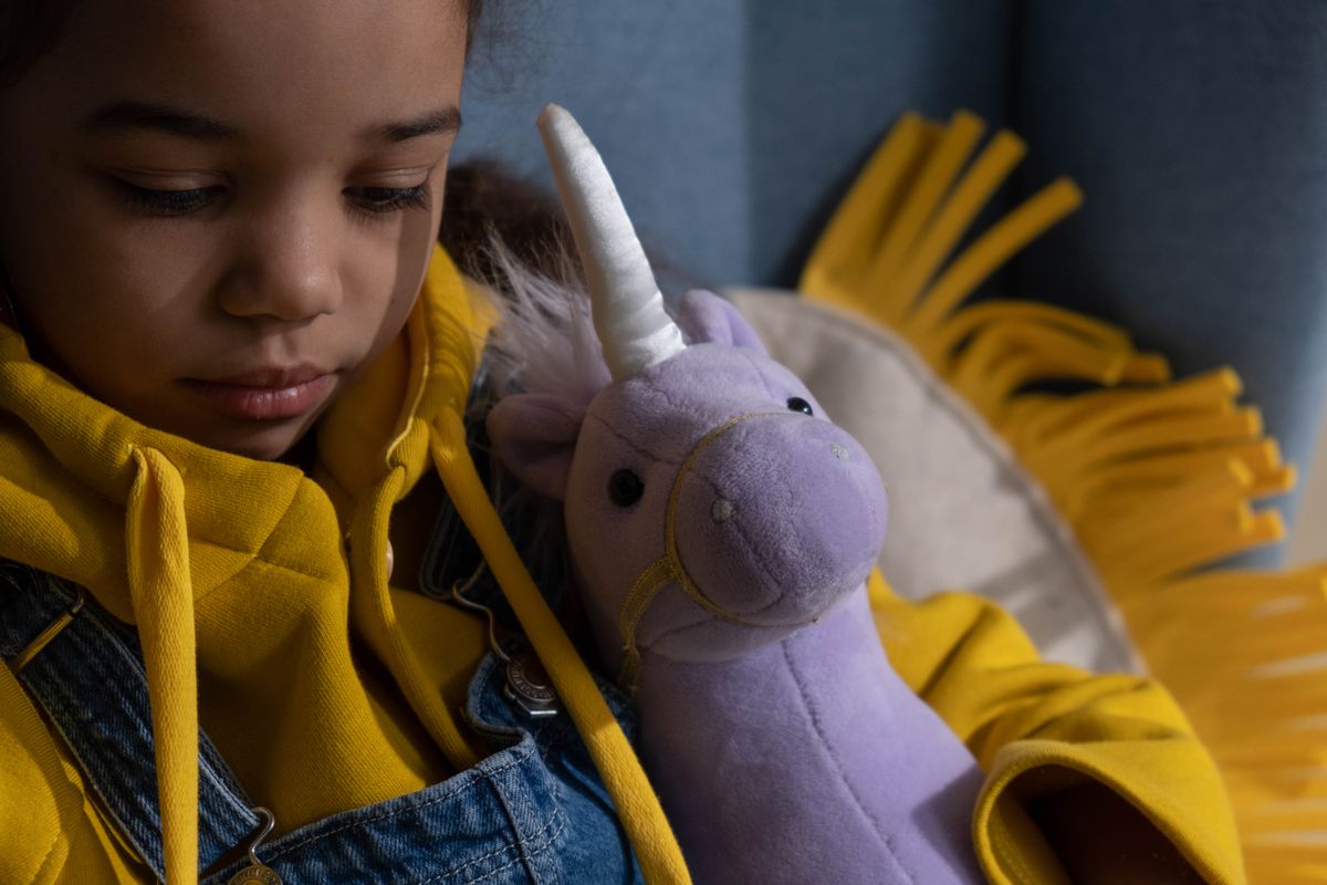 A young girl in a yellow hoodie holds a purple unicorn plush toy while sitting indoors.