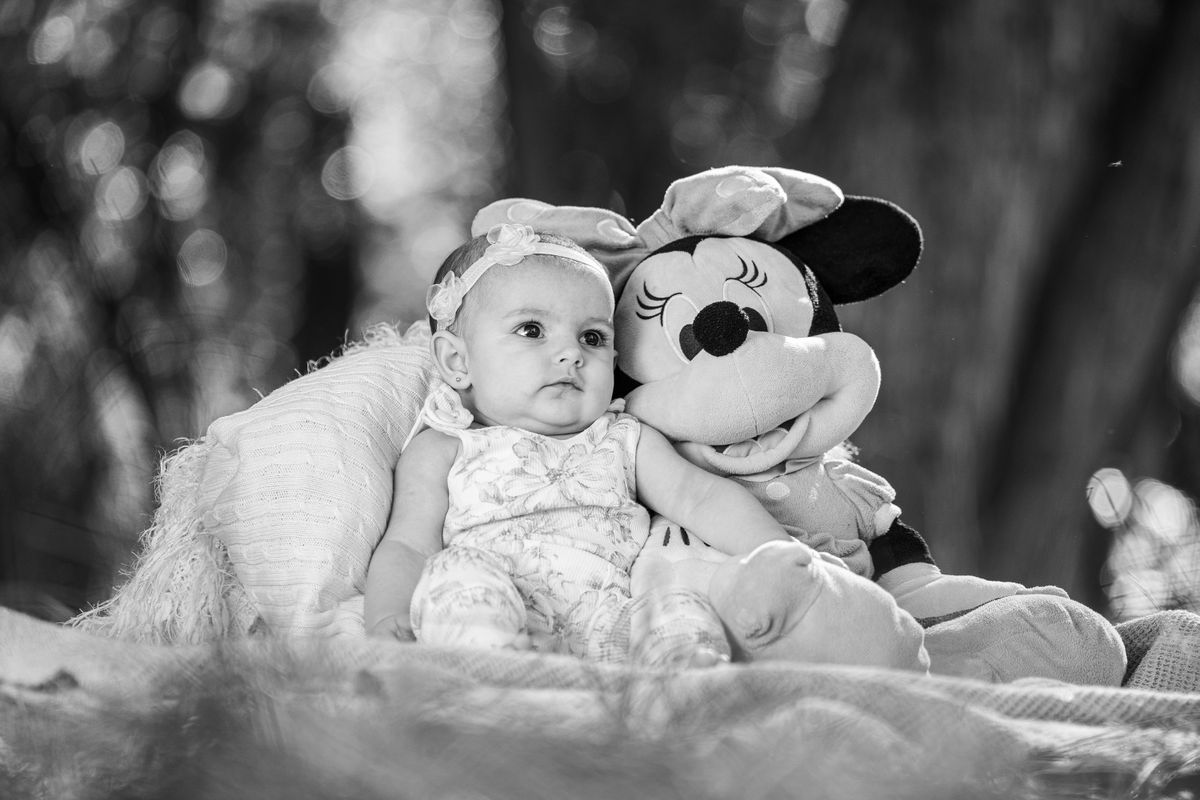 A cute baby girl sits outdoors with a Minnie Mouse plush in a black and white photograph.