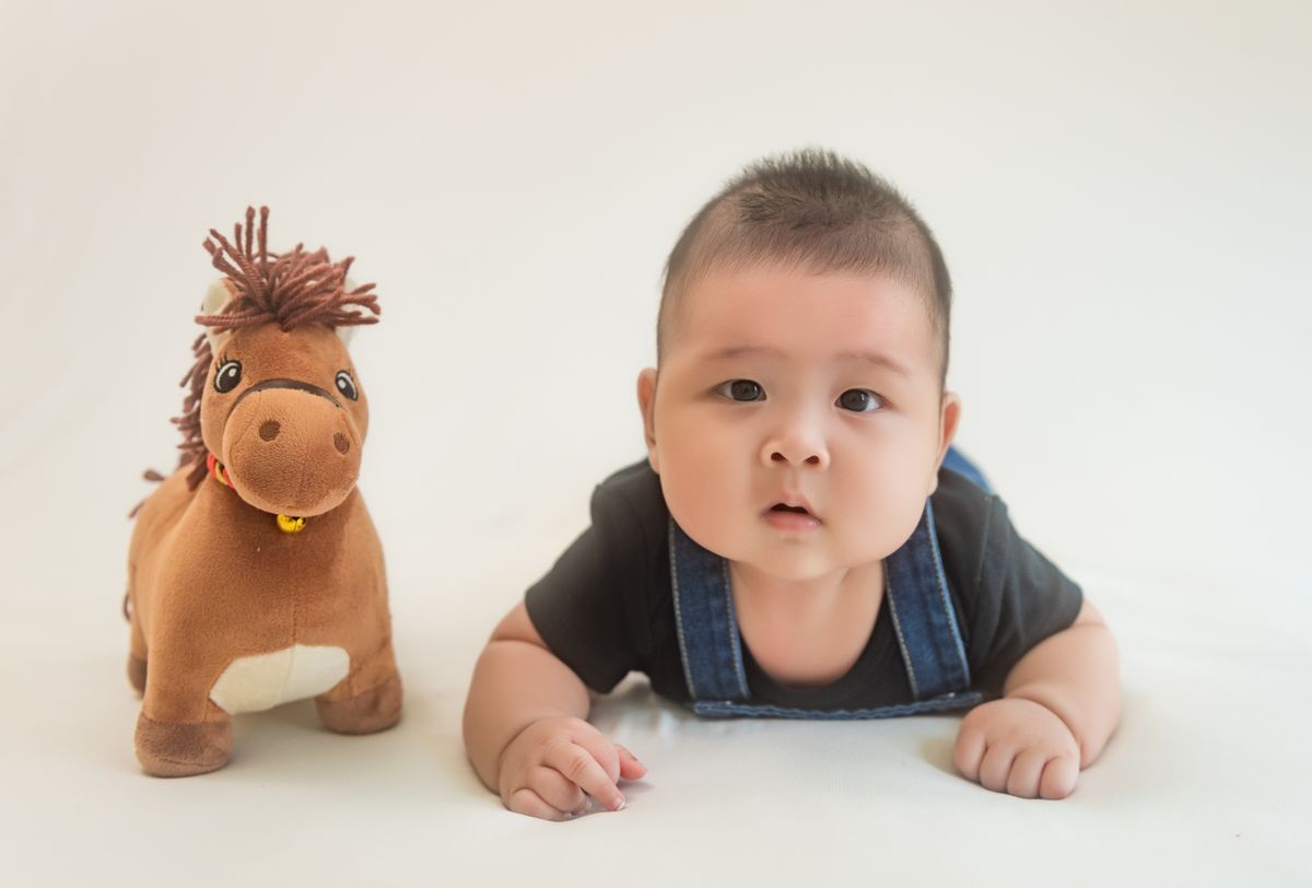 Cute baby crawling next to a plush horse toy on a white backdrop.