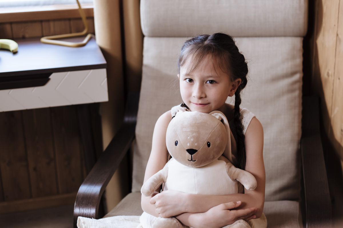 A cheerful girl with braided hair sitting indoors holding a plush toy, enjoying a moment of happiness.