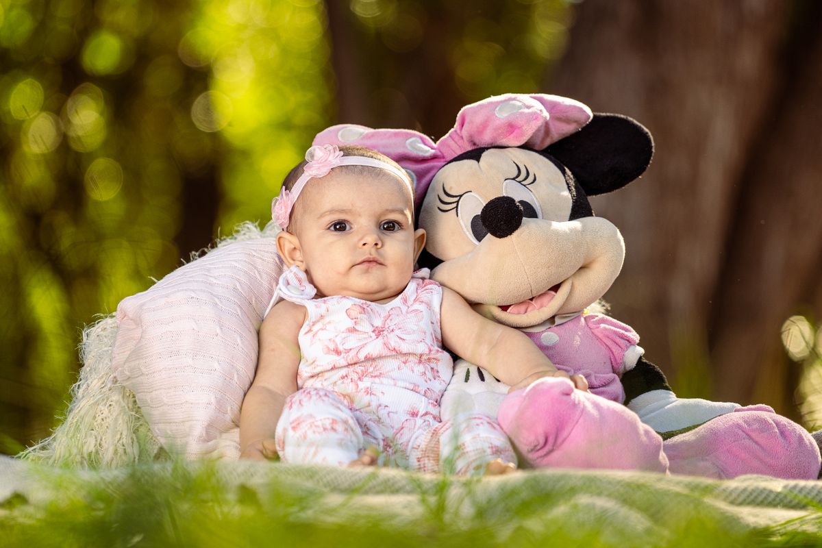 A cute baby sitting beside a Minnie Mouse plush toy in a sunny outdoor setting.