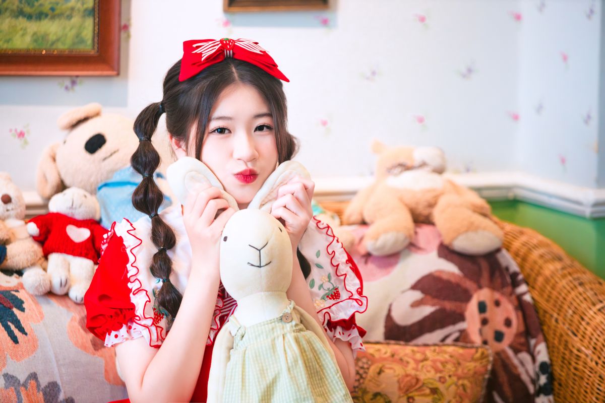 A cute child in a red dress cuddling a plush toy, surrounded by stuffed animals in a cozy, festive room.