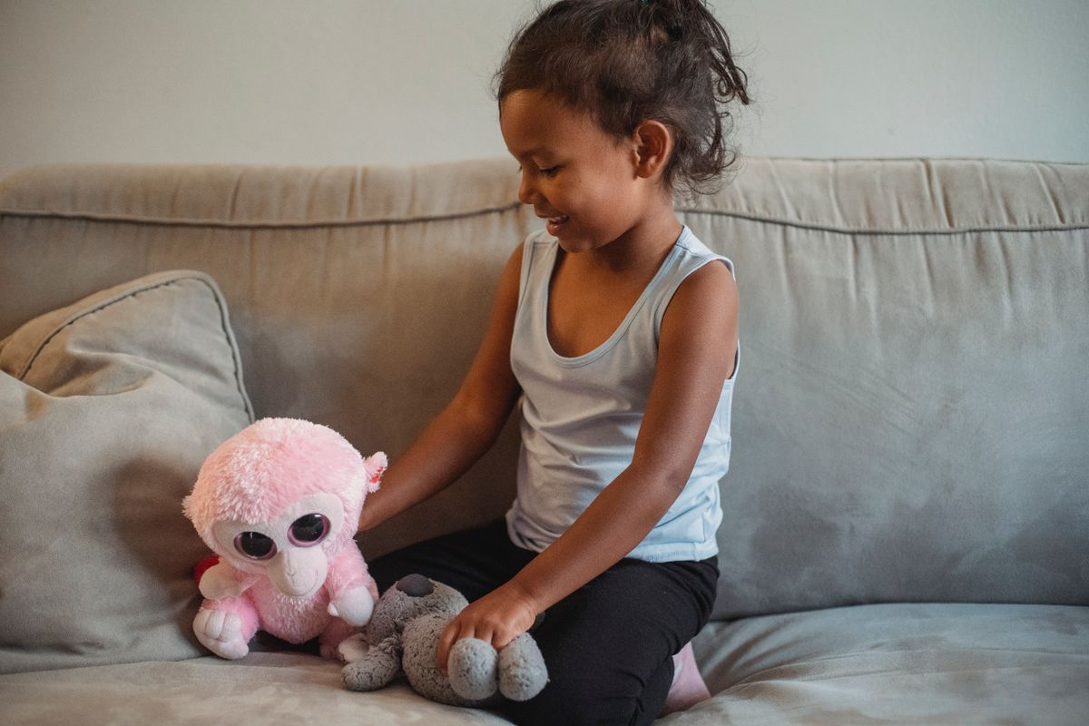 Smiling little Hispanic girl playing with soft toys on cozy sofa in living room