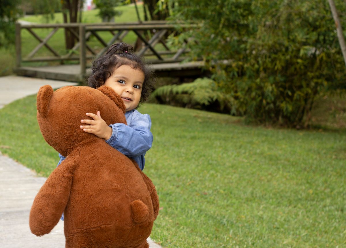 A cute child embraces a large plush teddy bear while standing on a grassy path.