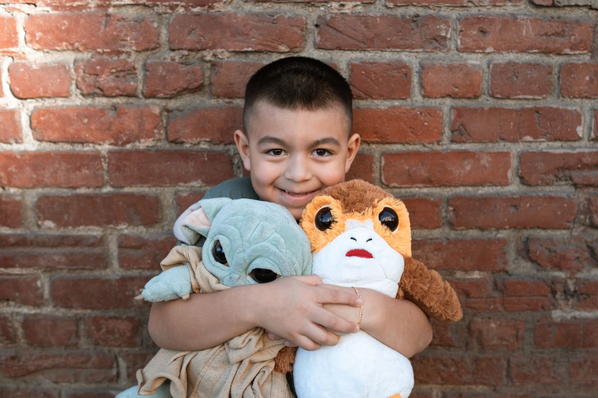 A cheerful child hugs Grogu and Porg plush toys against a rustic brick wall background.