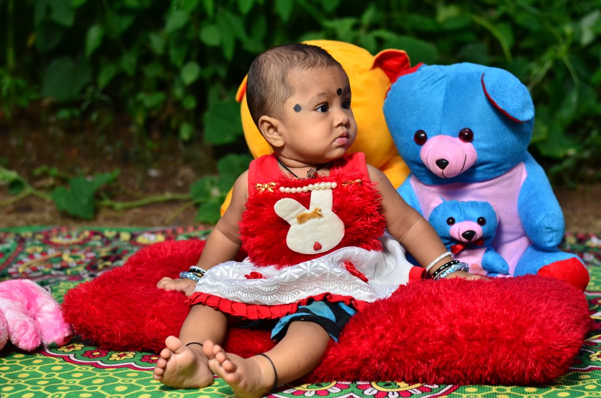 A cute baby sitting on a colorful blanket outdoors surrounded by plush toys.
