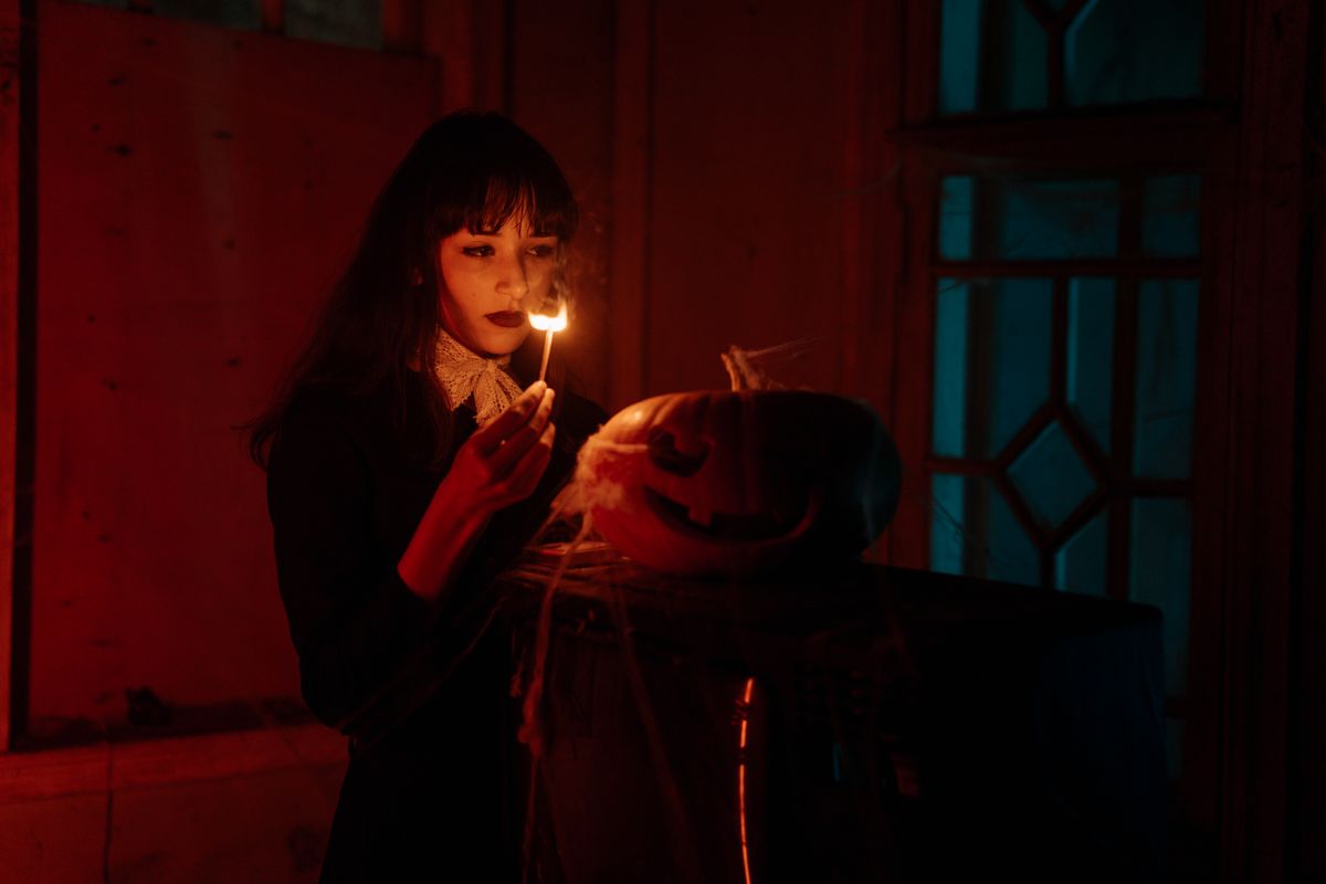 A woman lights a jack-o'-lantern with a match in a dimly lit, spooky setting.
