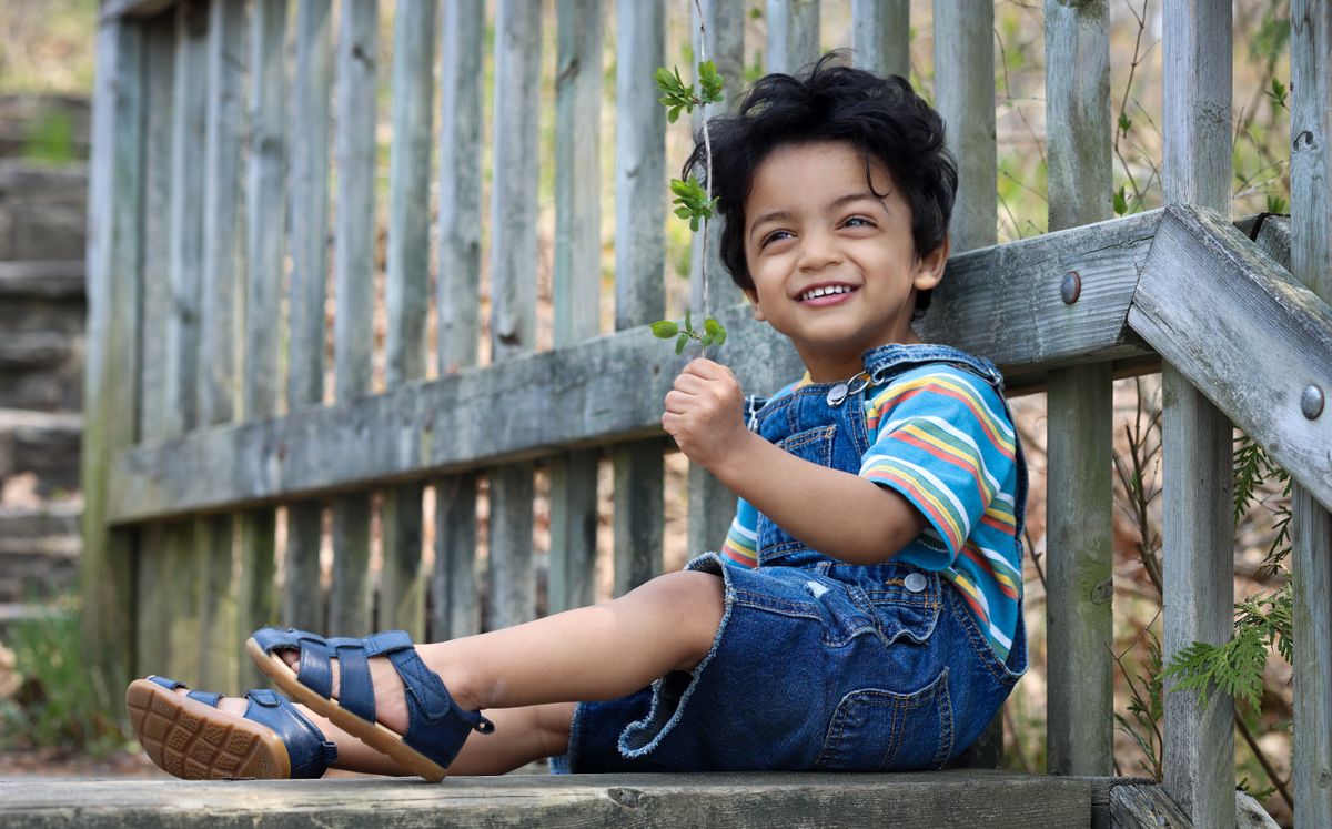 Smiling child in blue denim overalls sitting by a wooden fence on a sunny day.