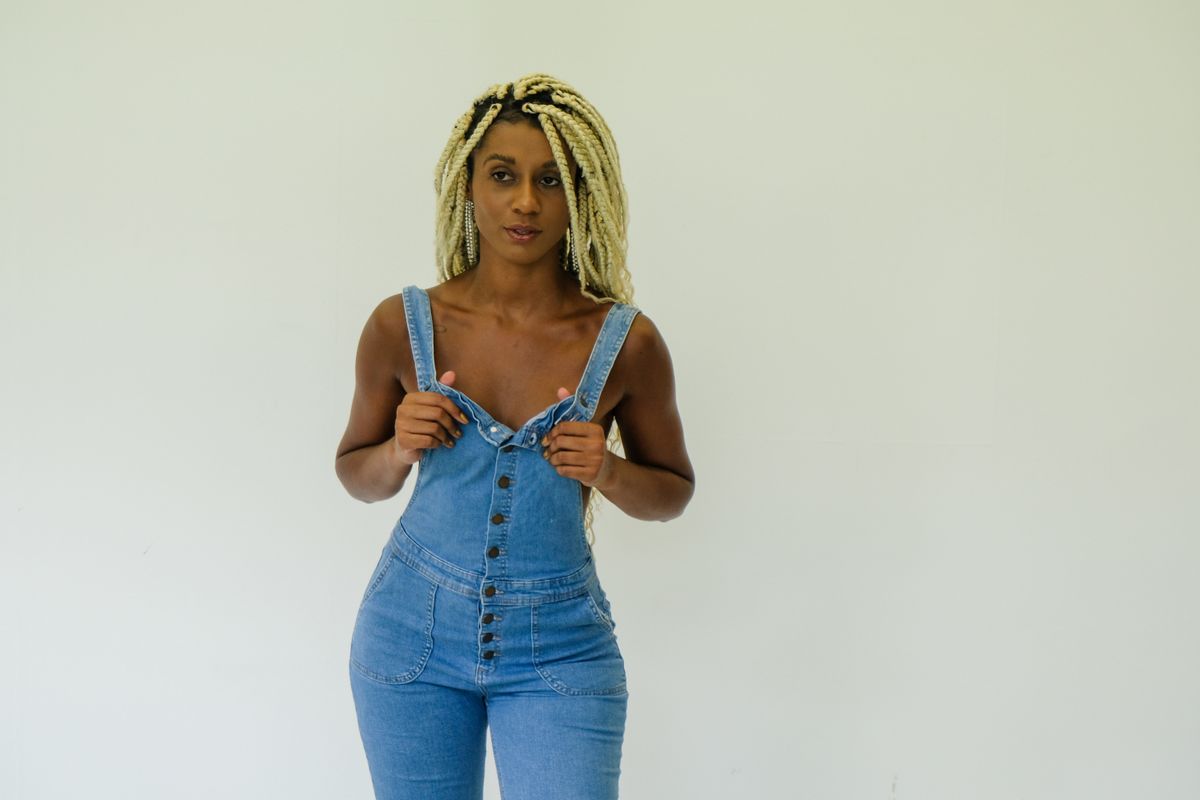 Fashion model with blonde braids in denim overalls standing against a white backdrop.