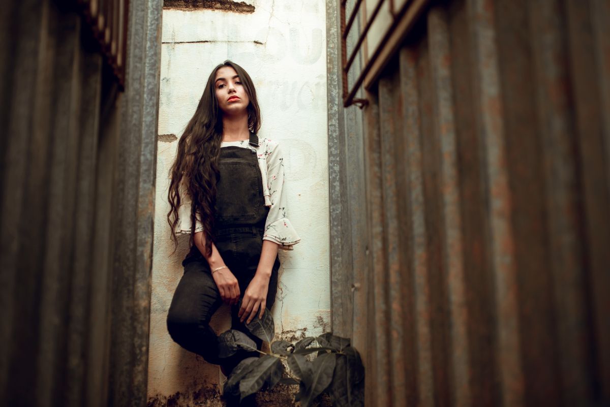 Fashionable young woman in urban alley, showcasing trendy style with worn overalls and flowing hair.