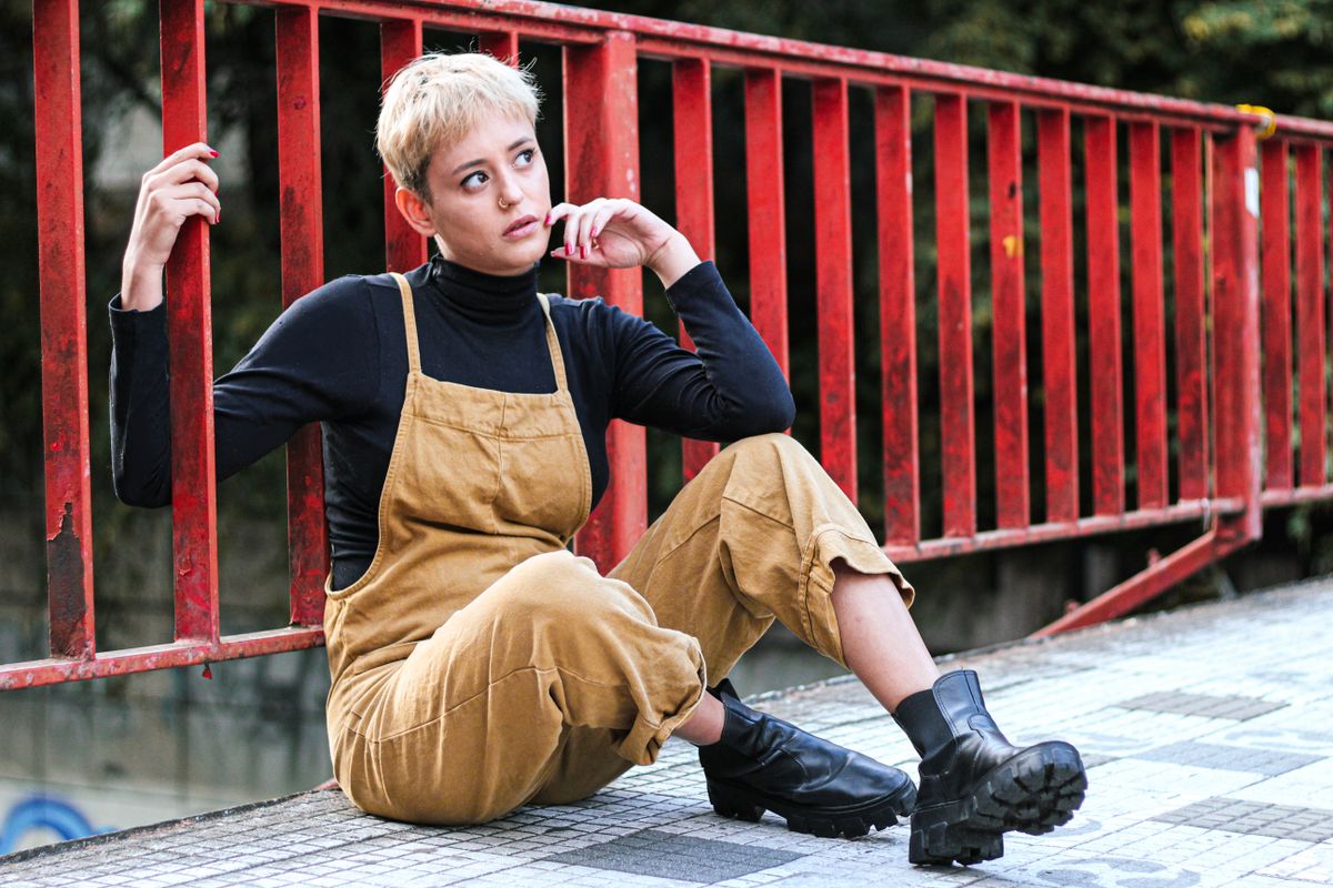 Fashionable woman in overalls sitting by a red fence, urban setting in São Paulo.