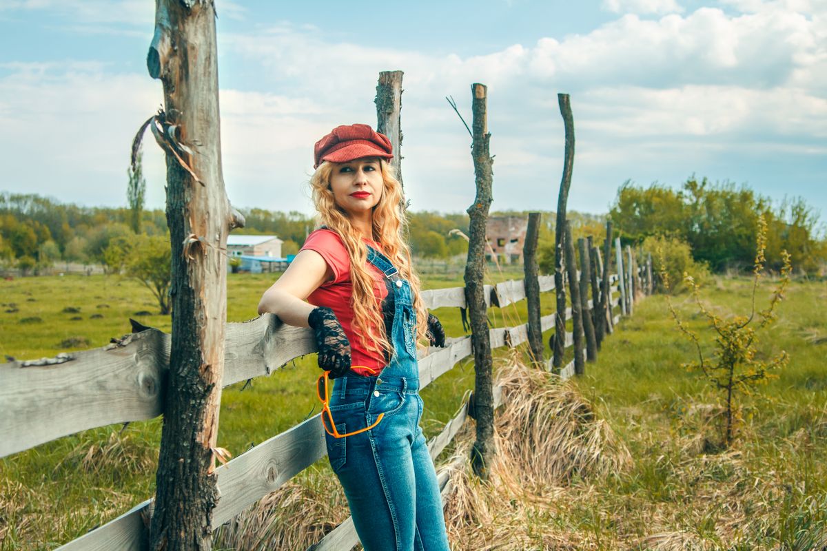 Charming blonde woman in a red cap and overalls relaxing by a rustic fence outdoors.