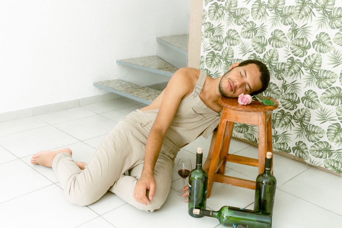 A man in overalls resting on a stool, surrounded by wine bottles indoors. Light and tranquil.