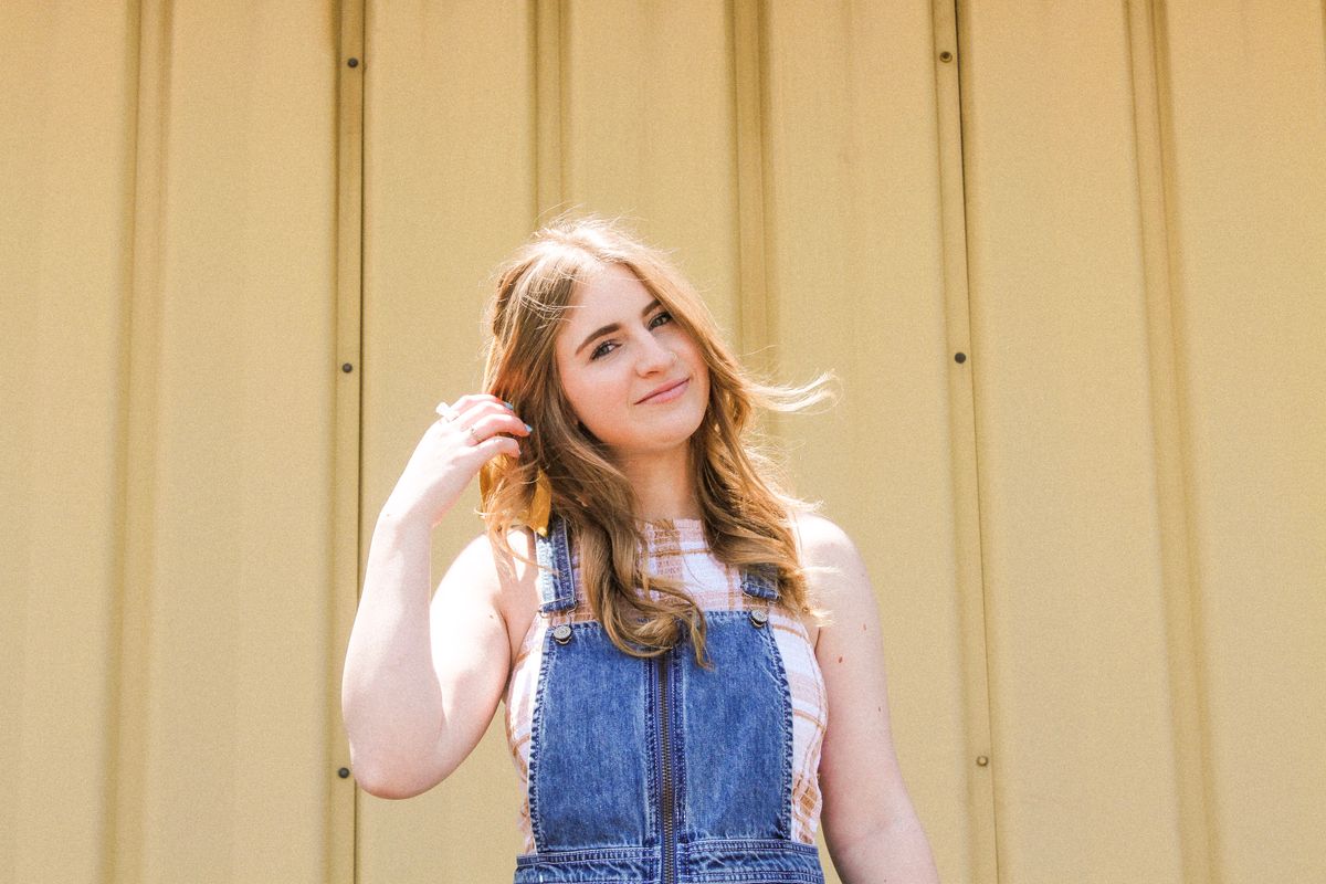 A young woman in denim overalls poses casually against a yellow background, smiling warmly.
