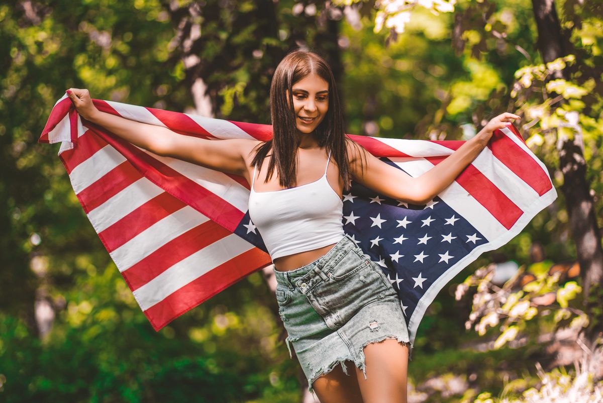 Young woman in a denim skirt holding the American flag outdoors in summer.