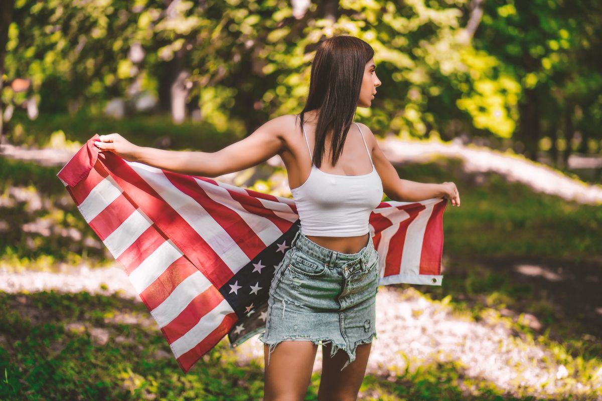 Young woman in crop top and denim skirt holding an American flag in a sunny forest.