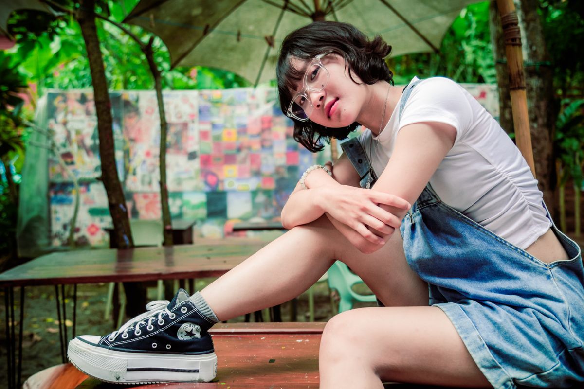 A trendy teenager wearing denim overalls poses casually at an outdoor cafe.