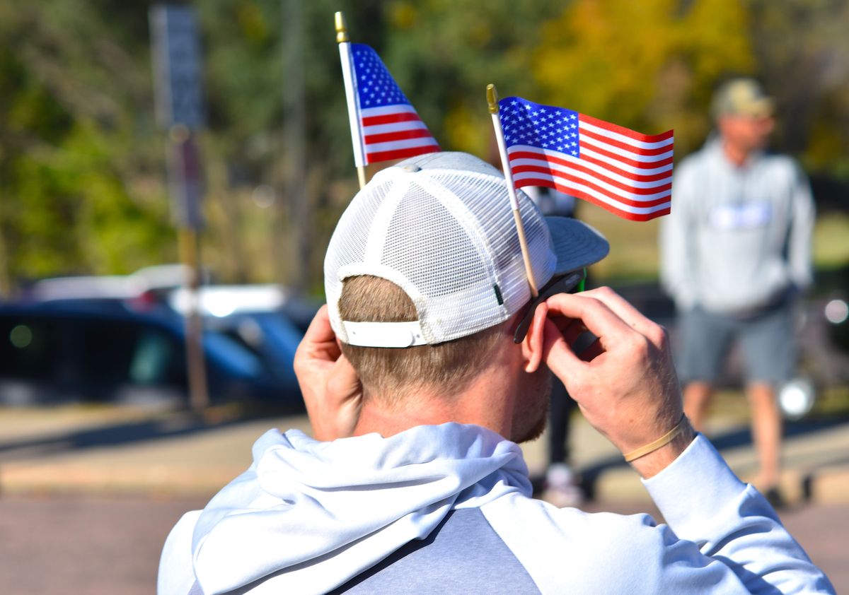 Rear view of a man wearing a hat adorned with small American flags at an outdoor event.