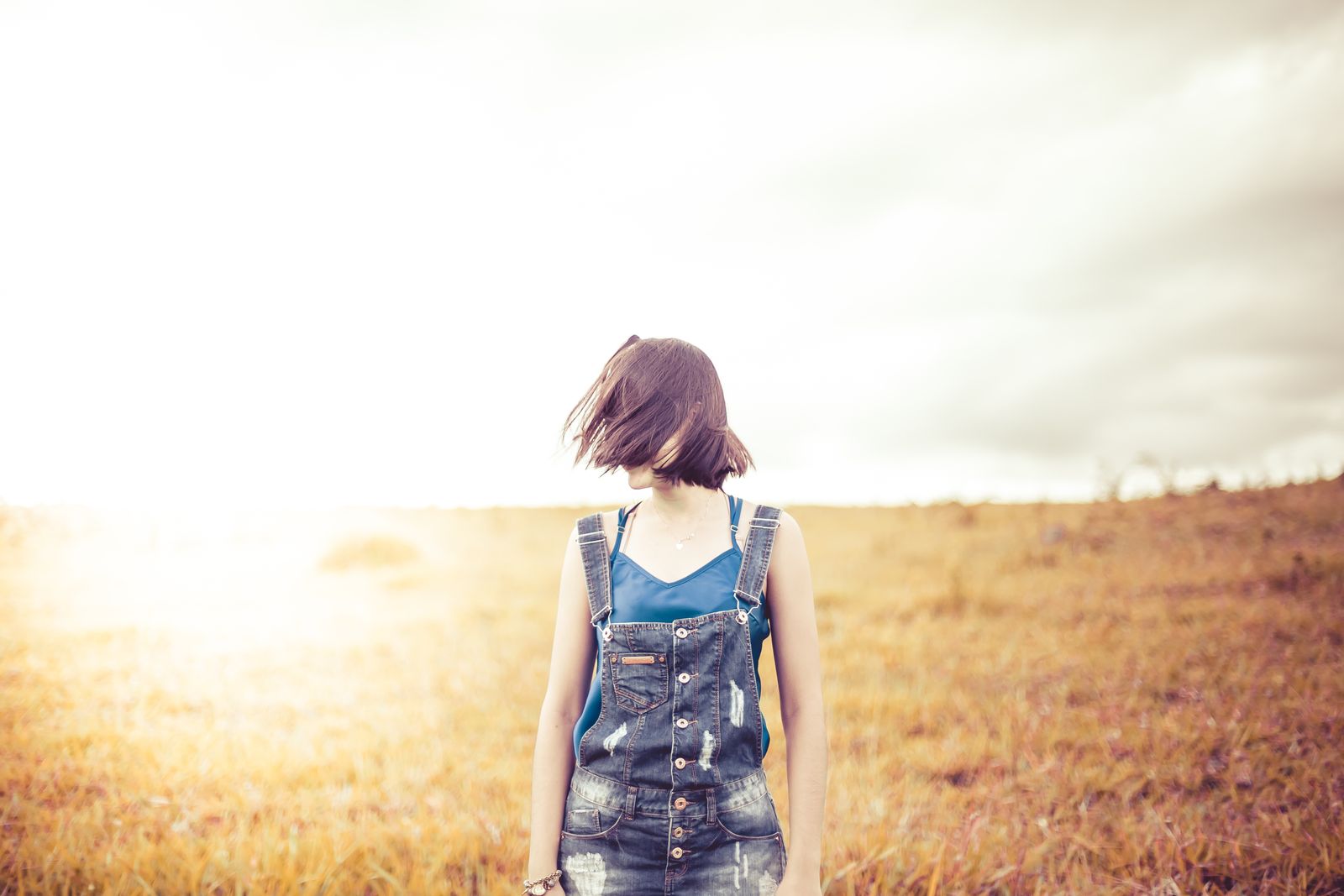 A woman stands in a sunlit field wearing denim overalls, embracing a carefree moment.