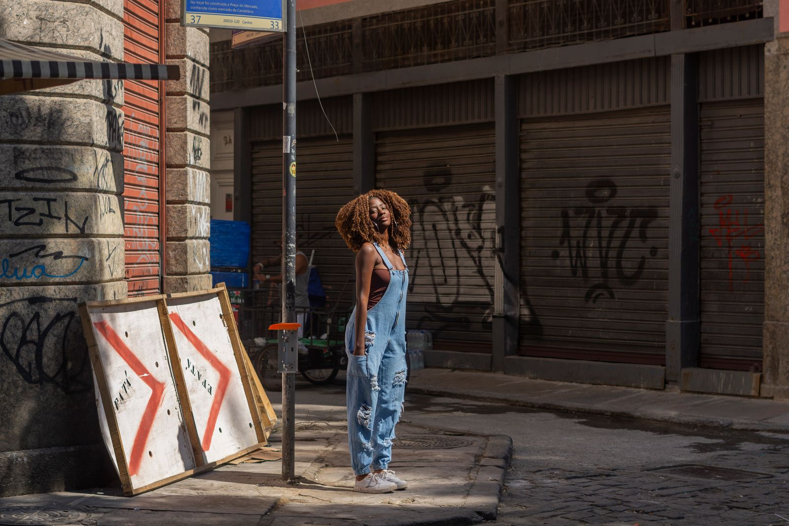 Woman standing confidently in urban street wearing denim overalls, surrounded by graffiti and city textures.