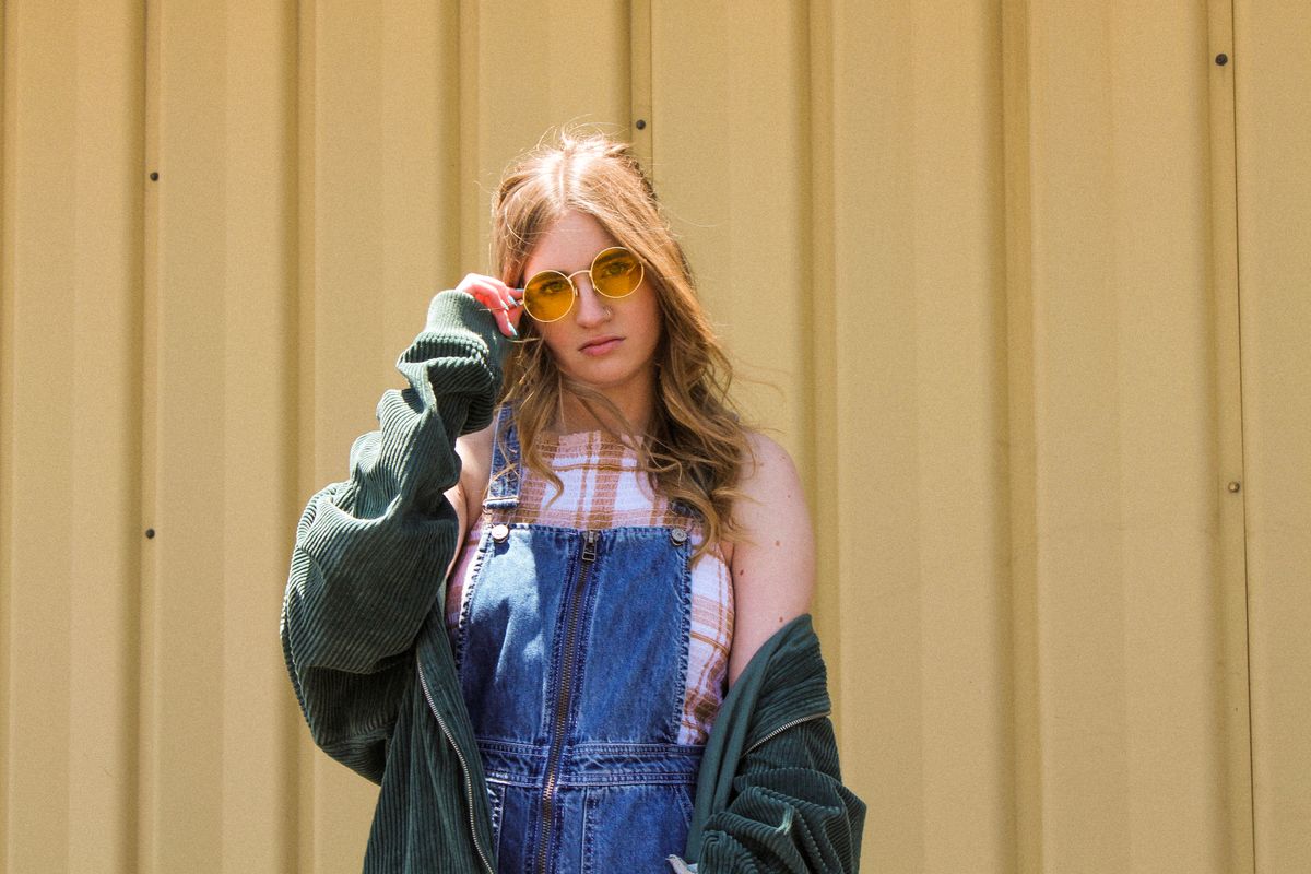 Portrait of a fashionable woman in denim dungarees wearing yellow sunglasses against a yellow wall.