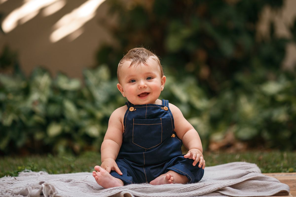 Adorable baby sitting outdoors on a blanket, wearing blue denim overalls and smiling brightly.