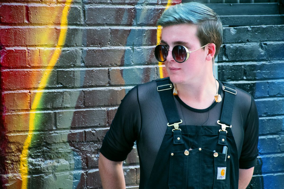 Trendy adult wearing sunglasses and overalls poses against a colorful graffiti wall, showcasing urban fashion.