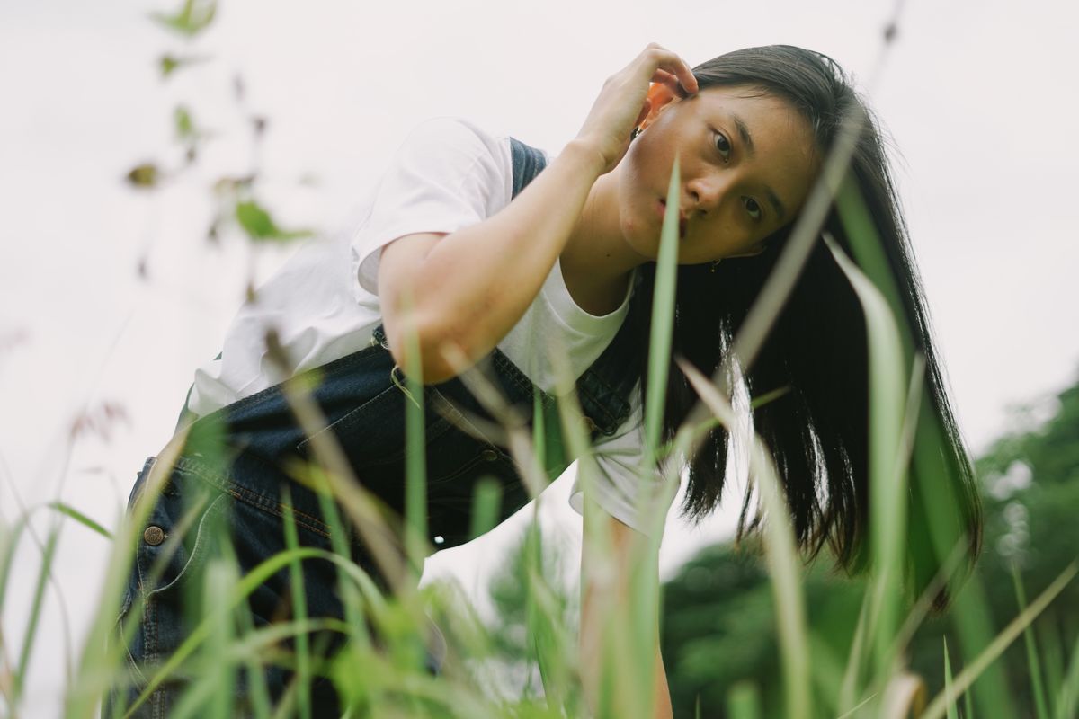 A young woman in a white shirt and denim overalls peers through tall grass outdoors.