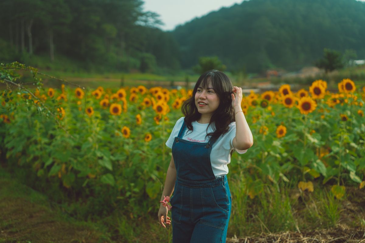 Young woman in denim overalls walking through vibrant sunflower fields.