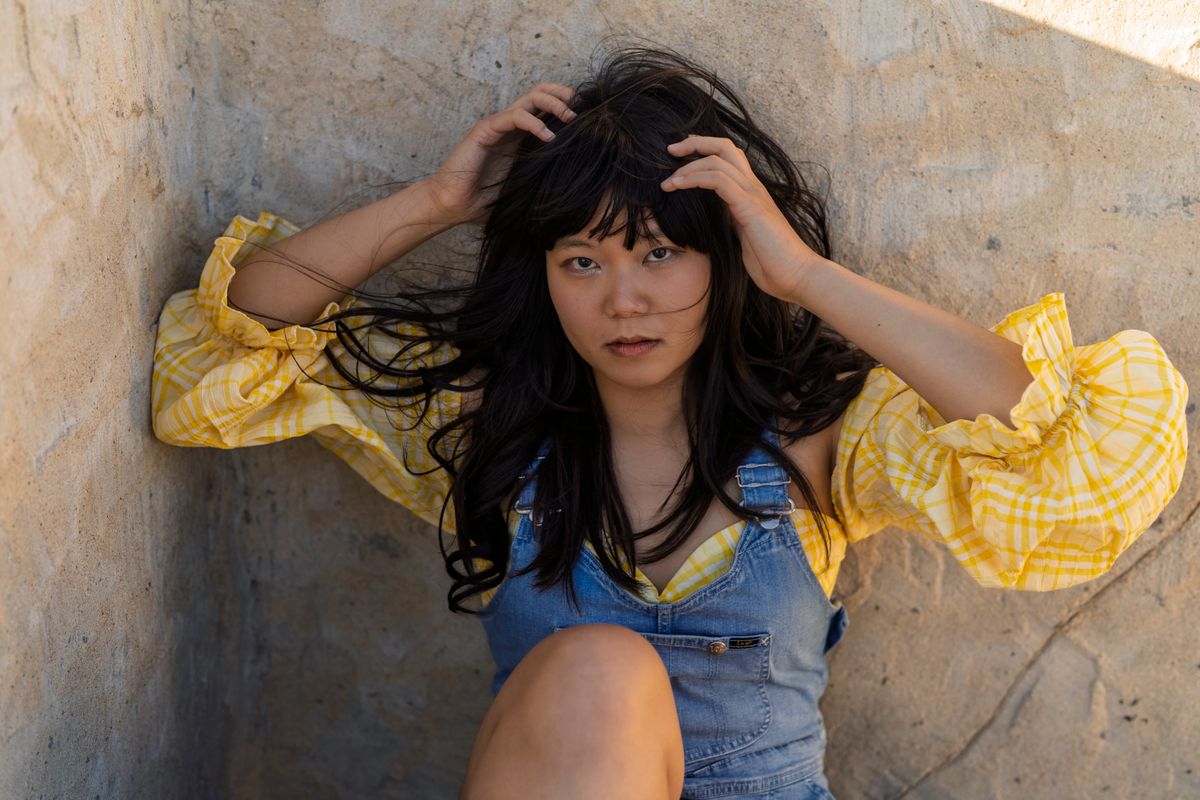 A young woman in a yellow plaid shirt and denim overalls sits thoughtfully against a concrete wall.