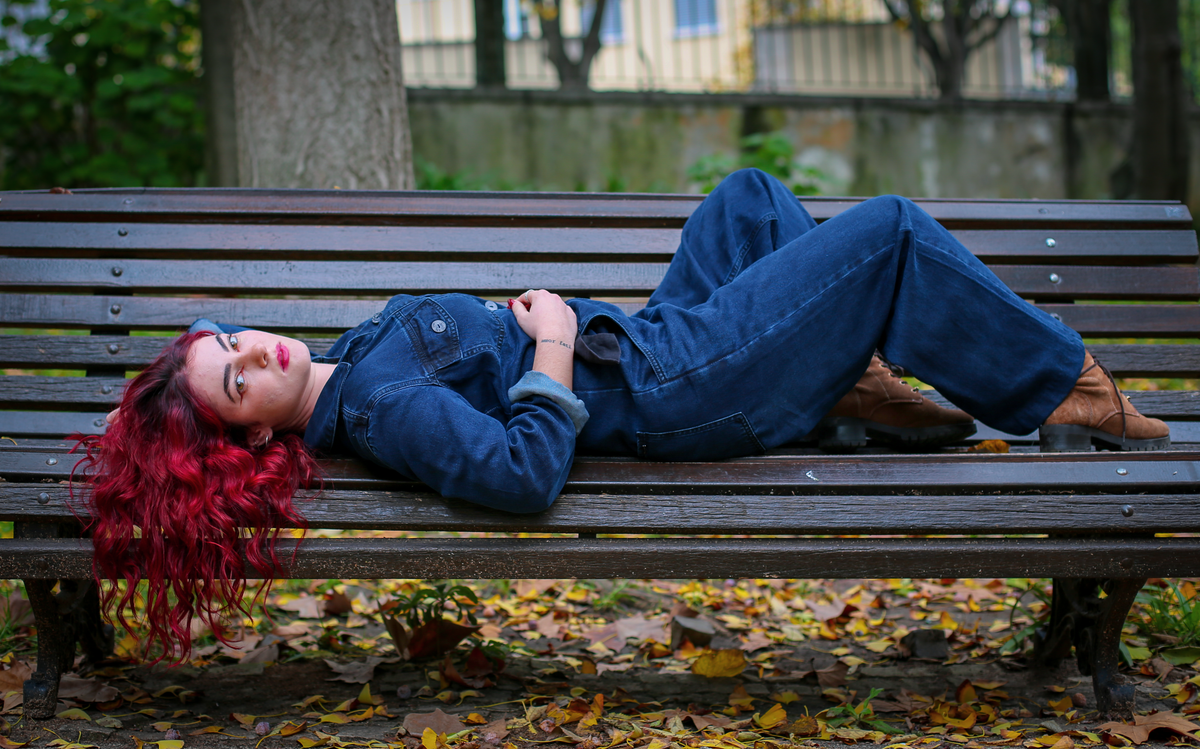 A woman with red hair lies on a park bench, surrounded by fall leaves, wearing denim.