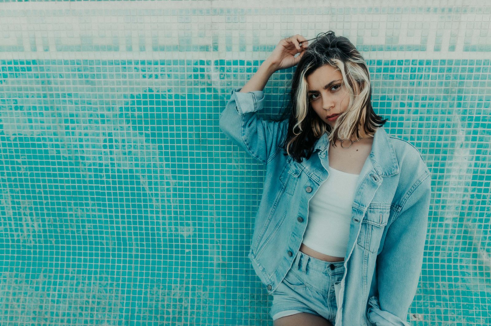 Stylish young woman in denim outfit posing against a blue mosaic wall, embodying modern fashion trends.