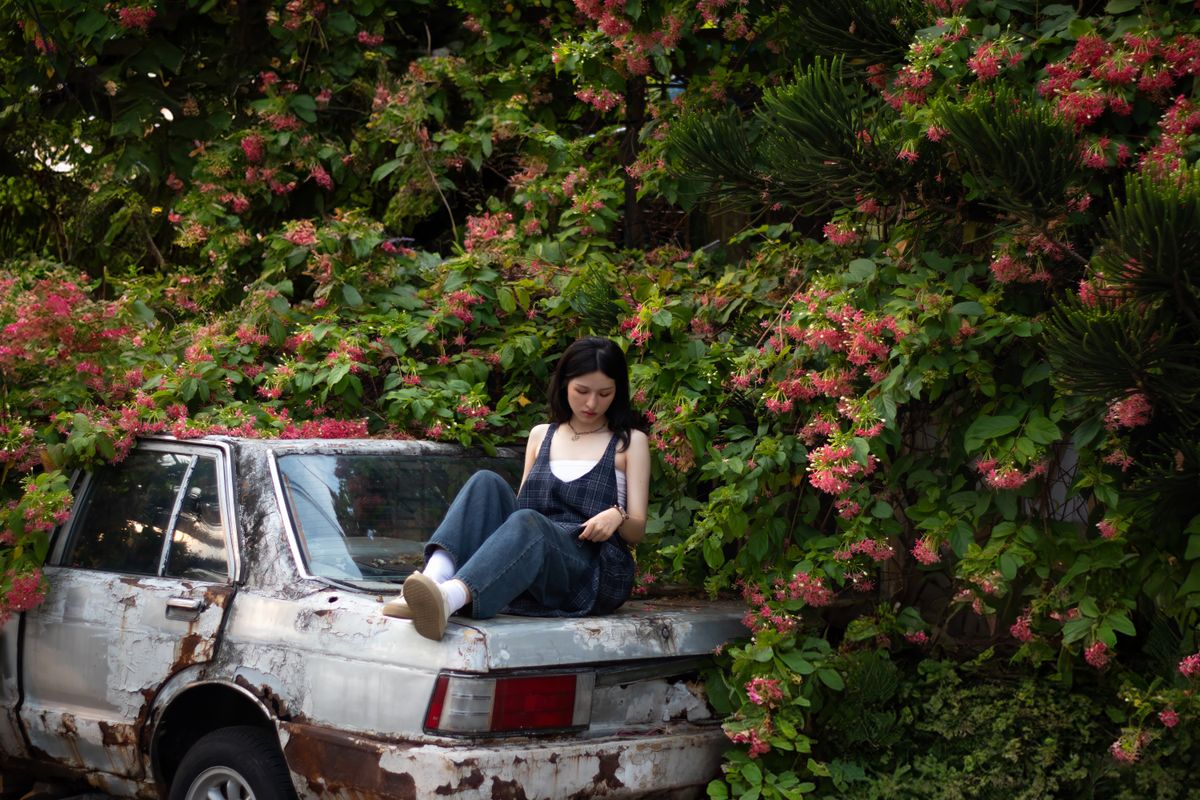 A woman in overalls sits on a rusty car, surrounded by lush greenery and pink flowers, creating a contrasting scene.
