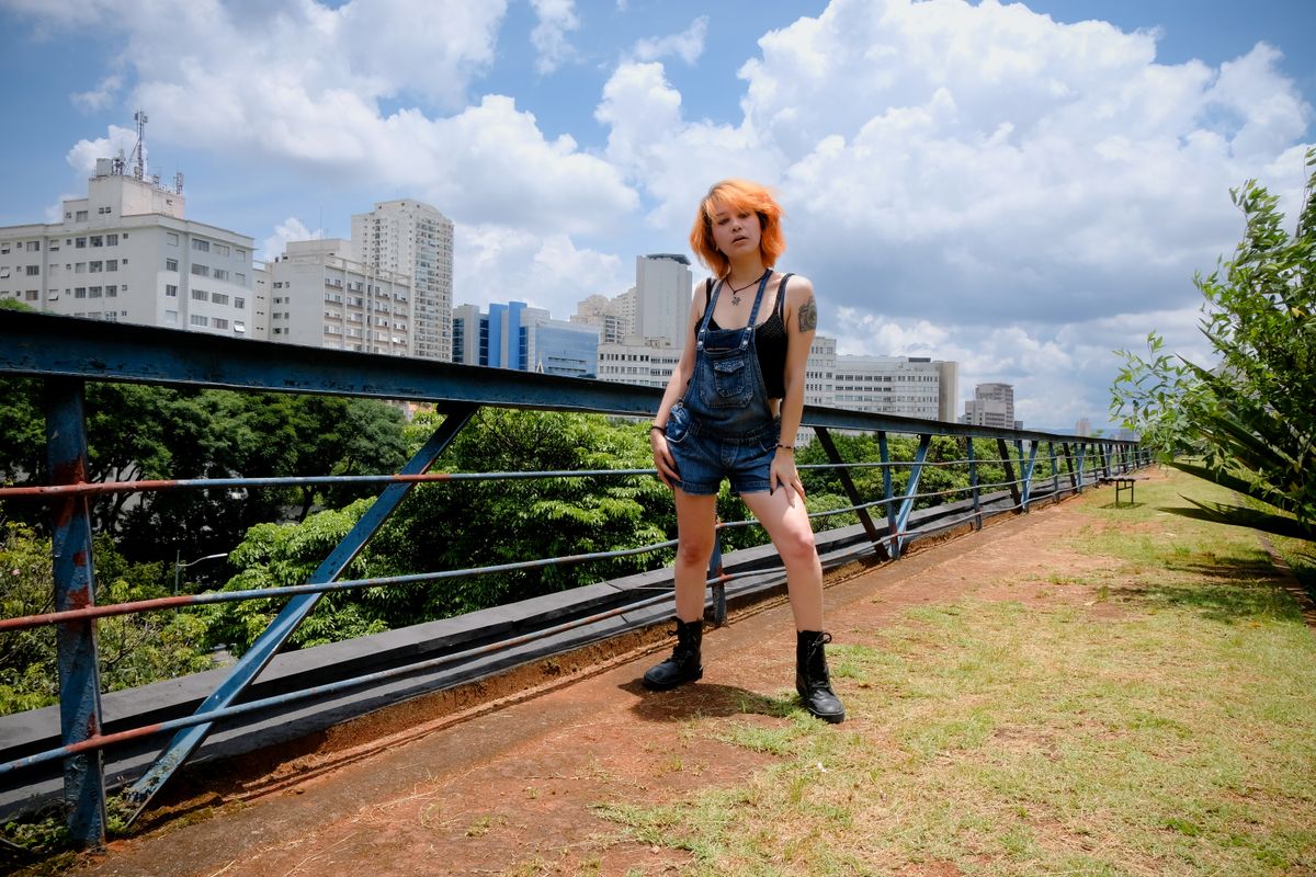 Young woman with red hair and tattoos in denim overalls standing outdoors in a São Paulo park.