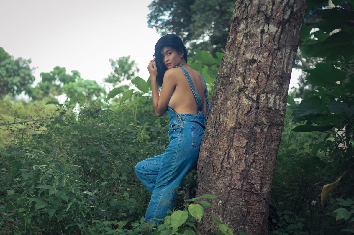 Asian woman in denim overalls leaning against a tree in a lush outdoor setting, capturing a serene moment.