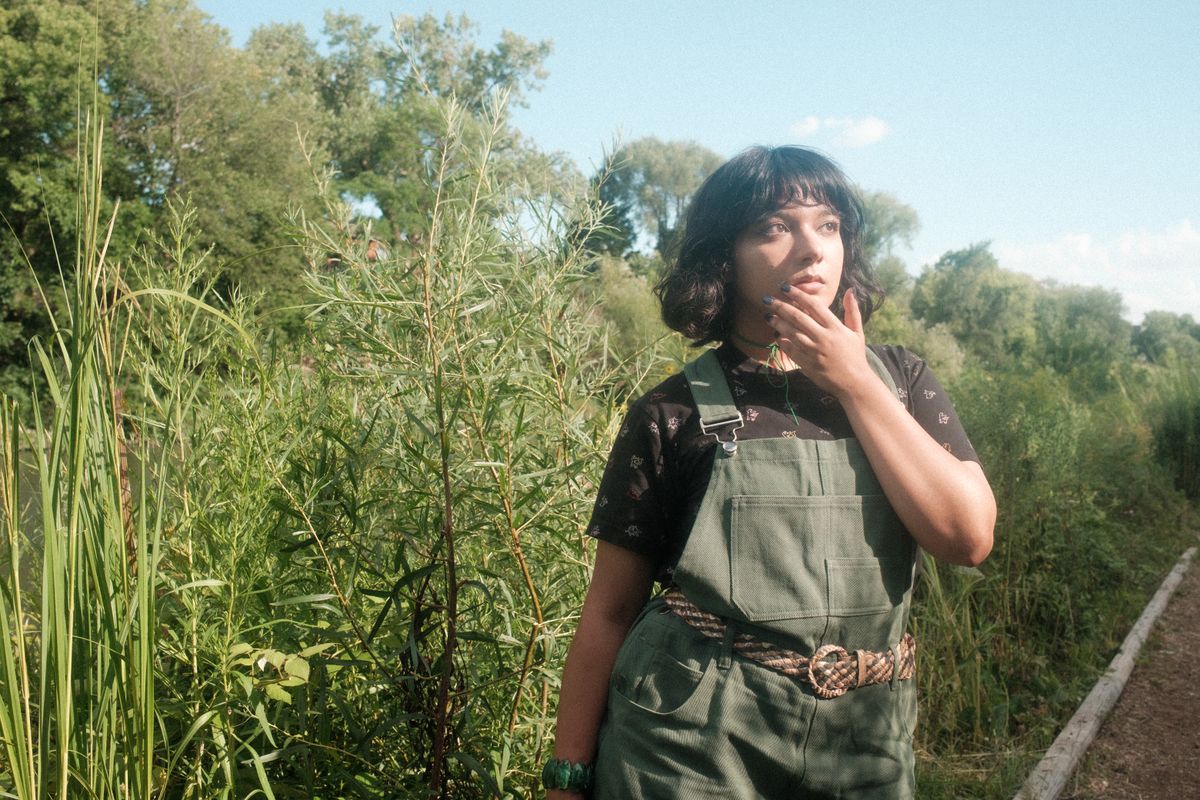 A young woman stands thoughtfully in lush greenery, wearing green overalls and a black top.