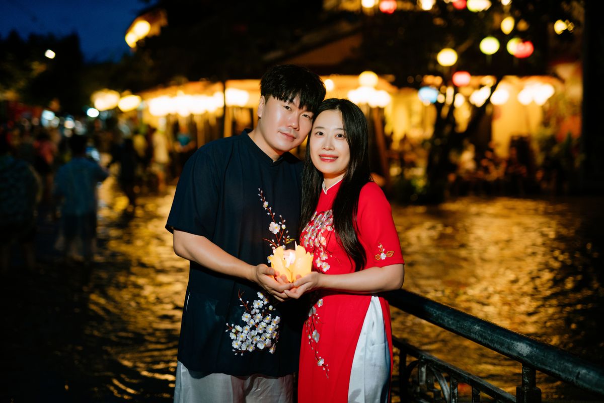 An Asian couple standing by a riverside, embracing and holding a lantern at night.