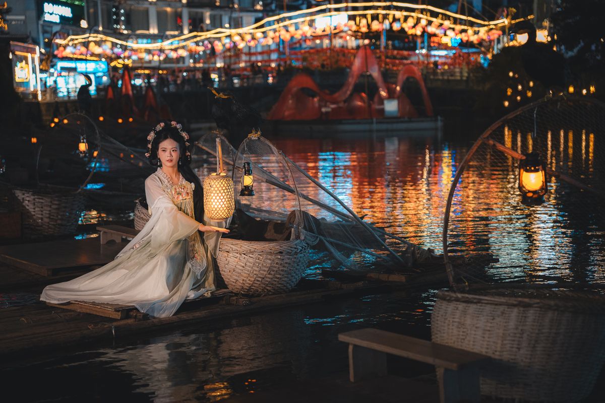 A woman in traditional Chinese attire holds a lantern on a raft at night in Guilin, China.